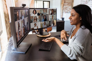 A woman in her home is on a zoom call with 18 other people on two screens.